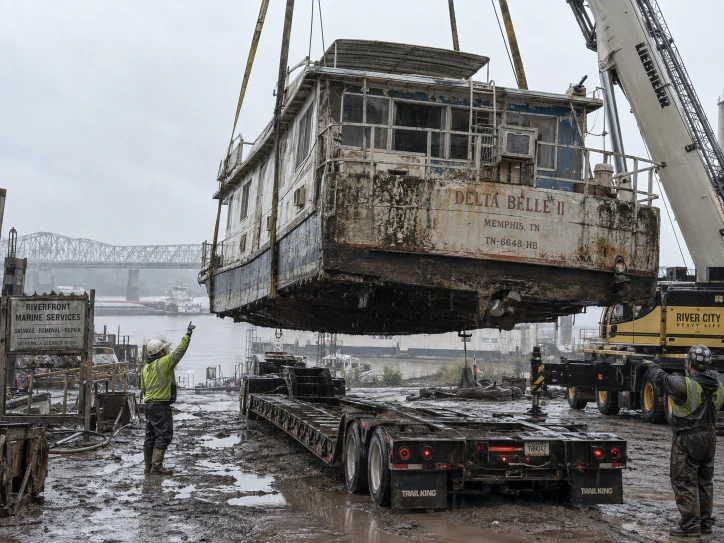 Weathered houseboat suspended above a flatbed beside a river and steel bridge